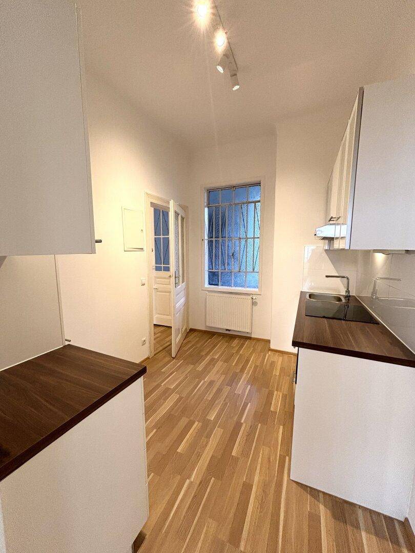 Leere Küche mit weißen Schränken, dunklen Arbeitsplatten und Fenster mit Gitter.