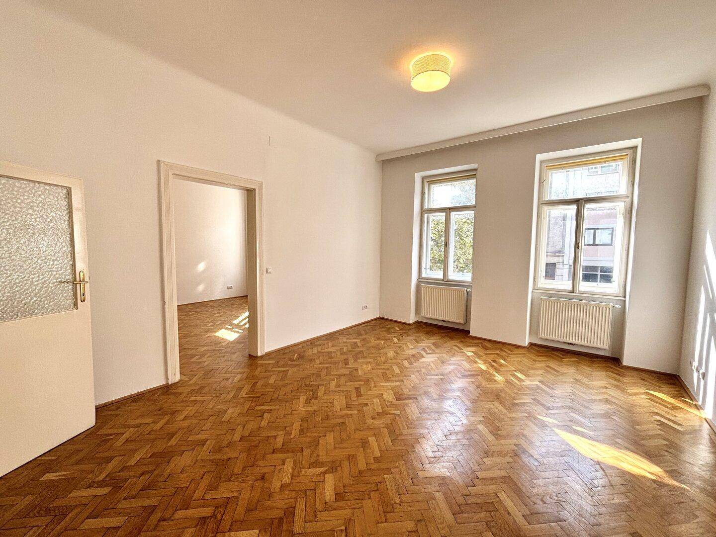 Leeres Wohnzimmer mit Fischgrätparkett, hohen Decken und Blick auf die Straße.