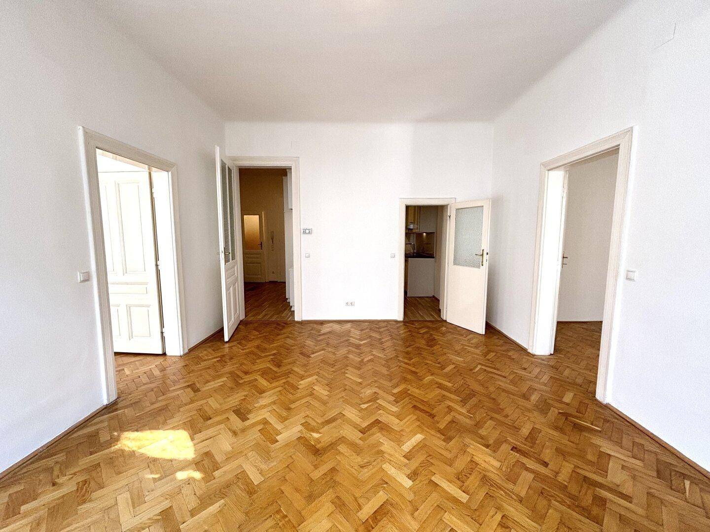 Großes, leeres Zimmer mit Fischgrätparkett und mehreren Türen zu angrenzenden Räumen.