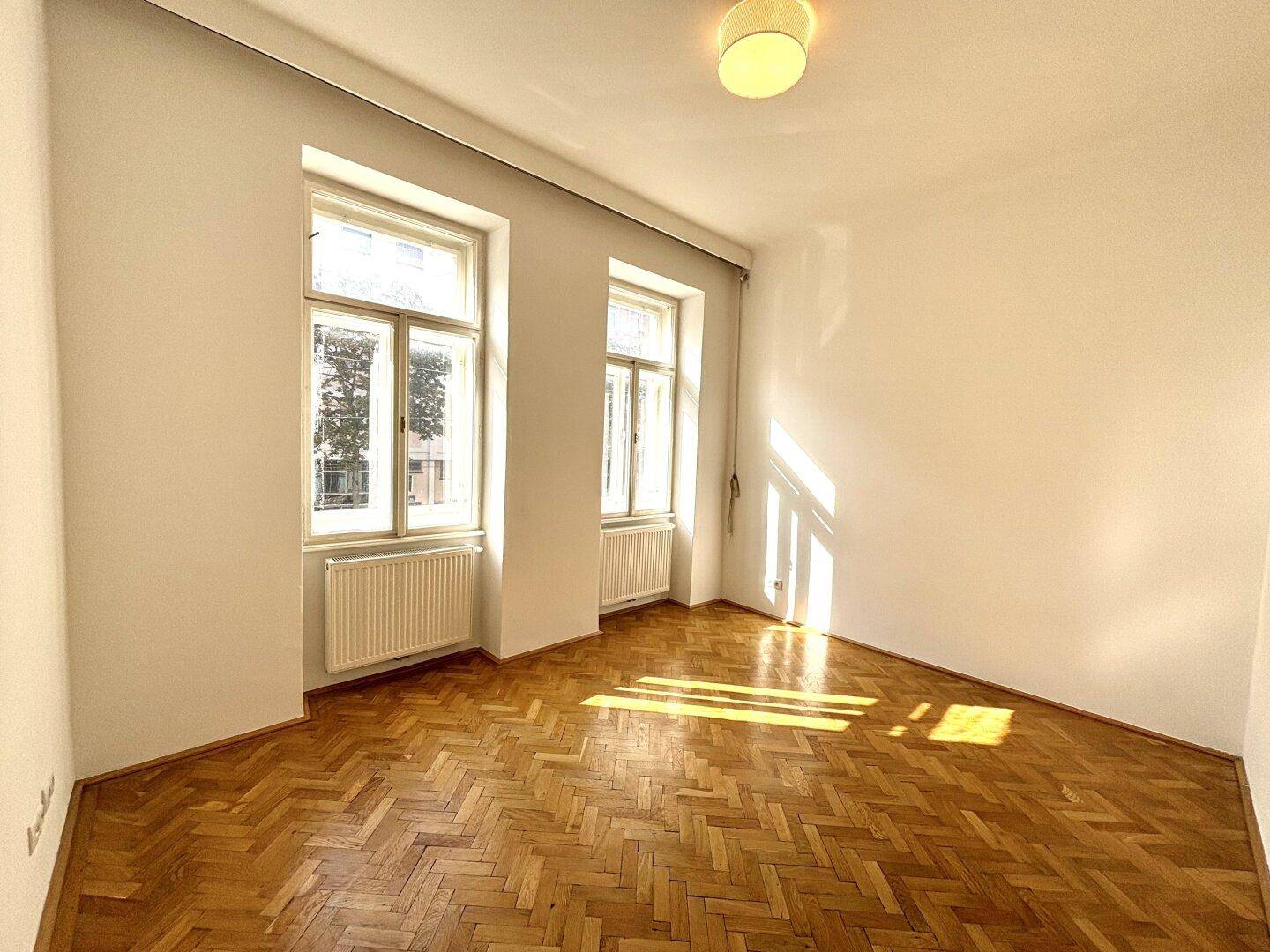 Geräumiges, leeres Zimmer mit Fischgrätparkett und viel Tageslicht durch große Fenster.