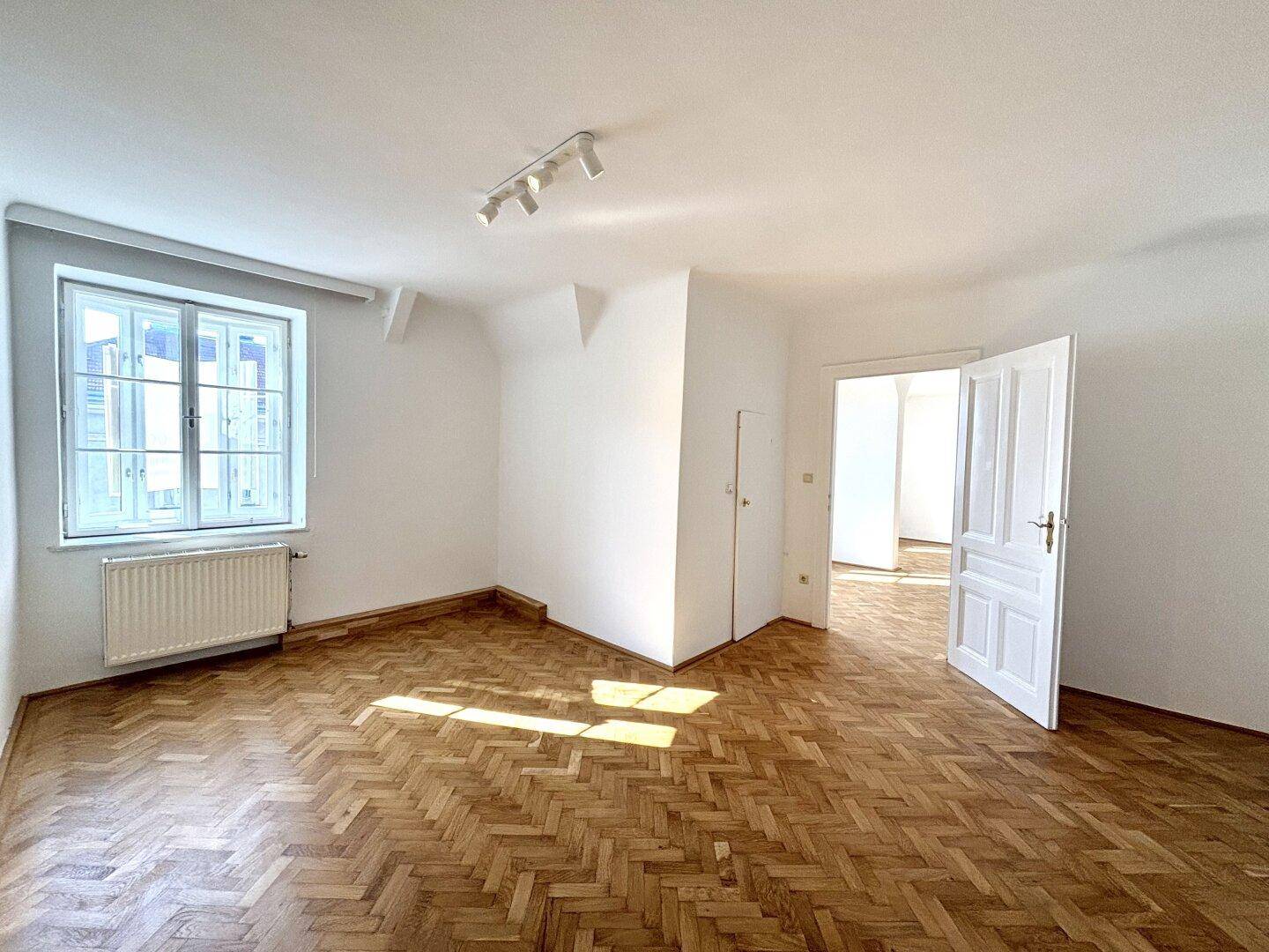 Leeres Schlafzimmer mit Parkettboden, großen Fenstern und viel natürlichem Lichteinfall.