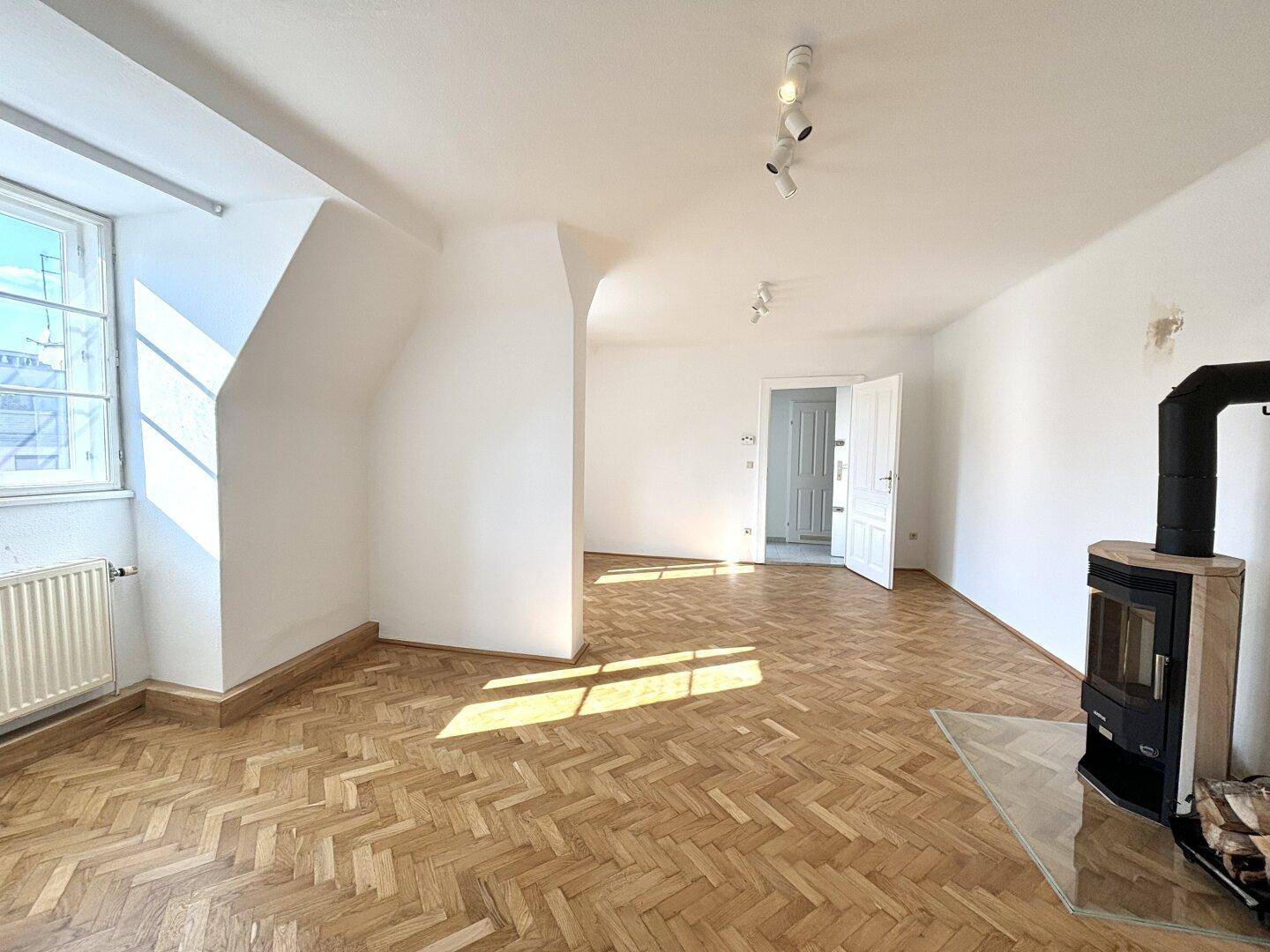 Geräumiger, leerer Wohnbereich mit Parkettboden, Kaminofen und sonnigem Fenster.