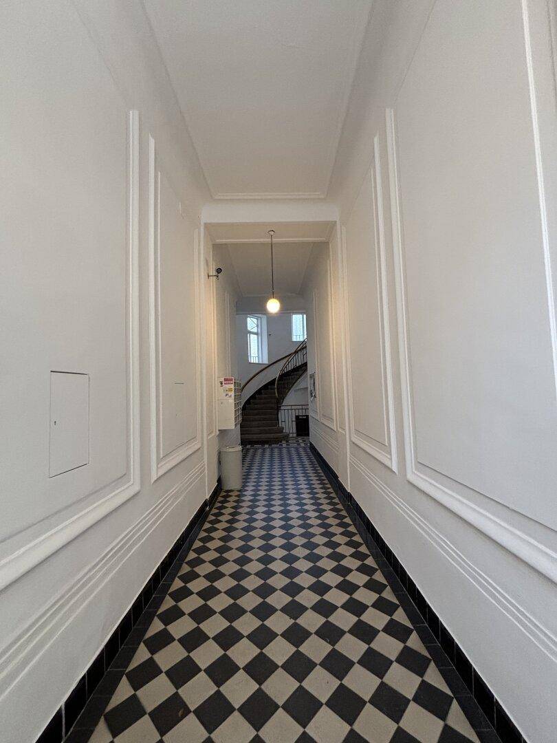 Langer Flur im Altbau mit Schachbrettmuster-Fliesen und eleganter Wendeltreppe im Hintergrund.
