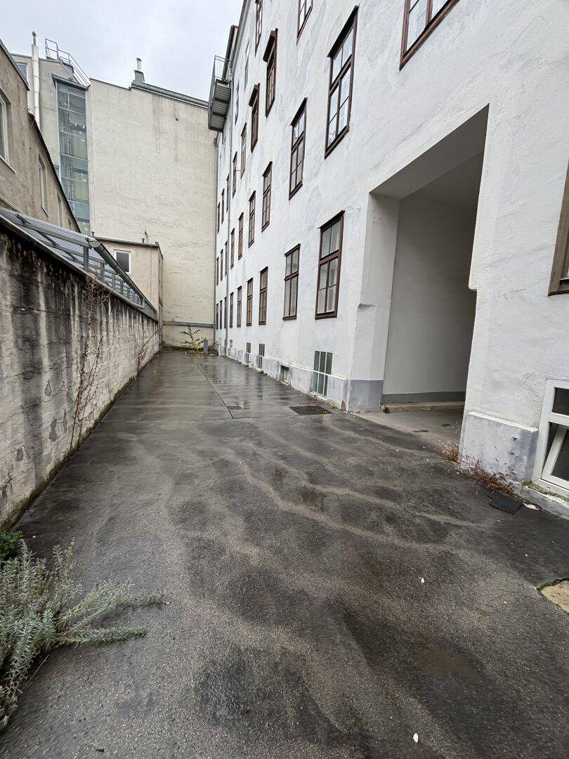 Außenansicht eines Gebäudes mit schmalem Durchgang, nasser Asphaltfläche und spärlicher Vegetation an der Wand.