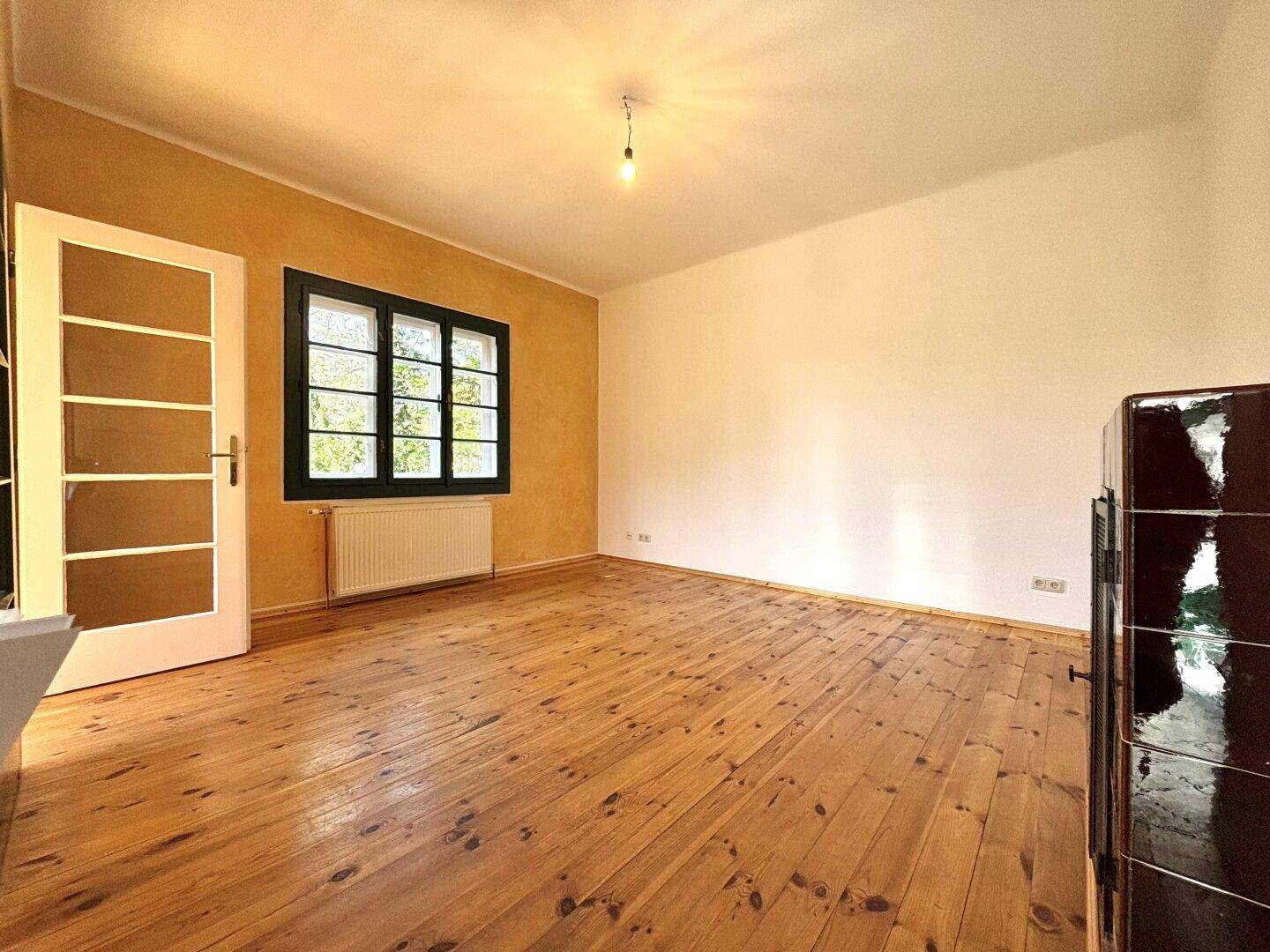 Helles Zimmer mit Holzdielenboden, gelber Akzentwand und einem traditionellen Kachelofen.