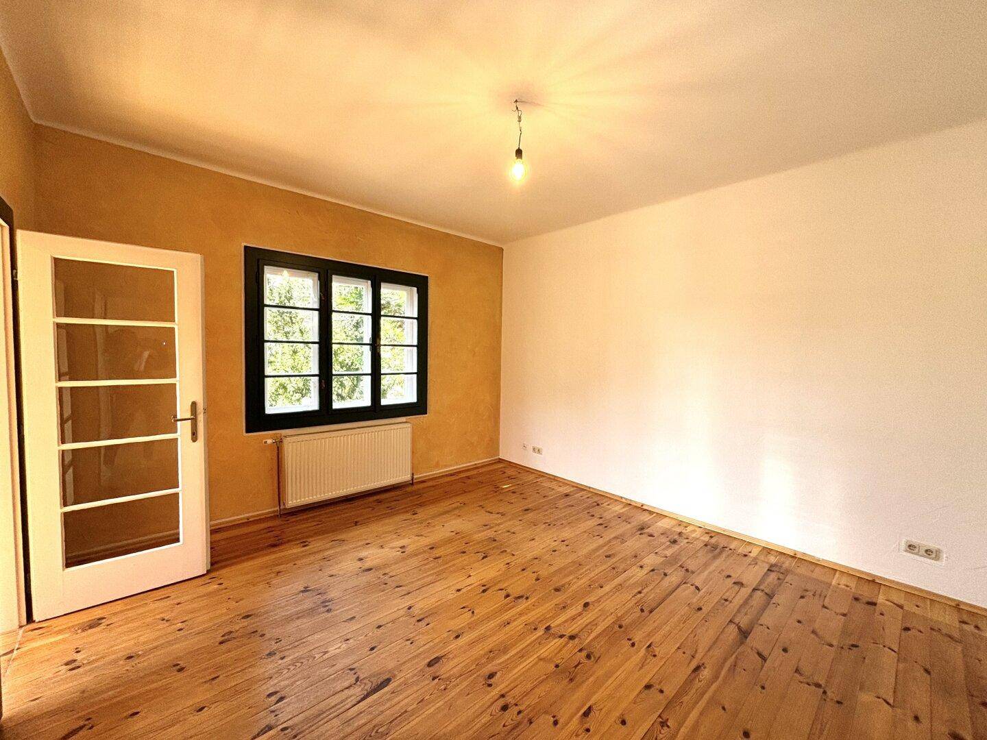 Geräumiges Zimmer mit Holzdielenboden, gelber Wand und einem Fenster mit Blick ins Grüne.
