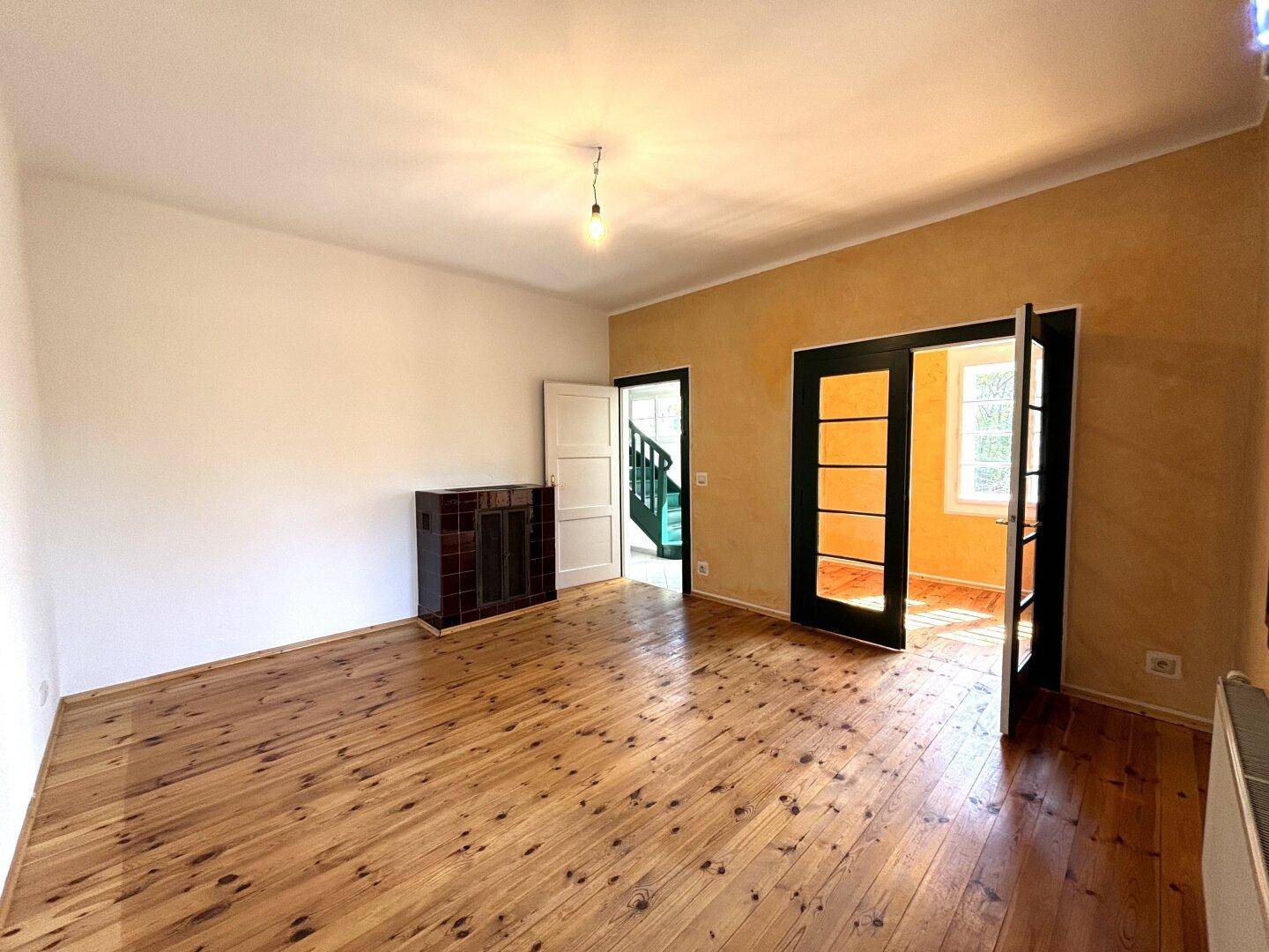 Wohnraum mit Holzdielenboden, Kachelofen und Zugang zu weiteren Zimmern und Treppenhaus.