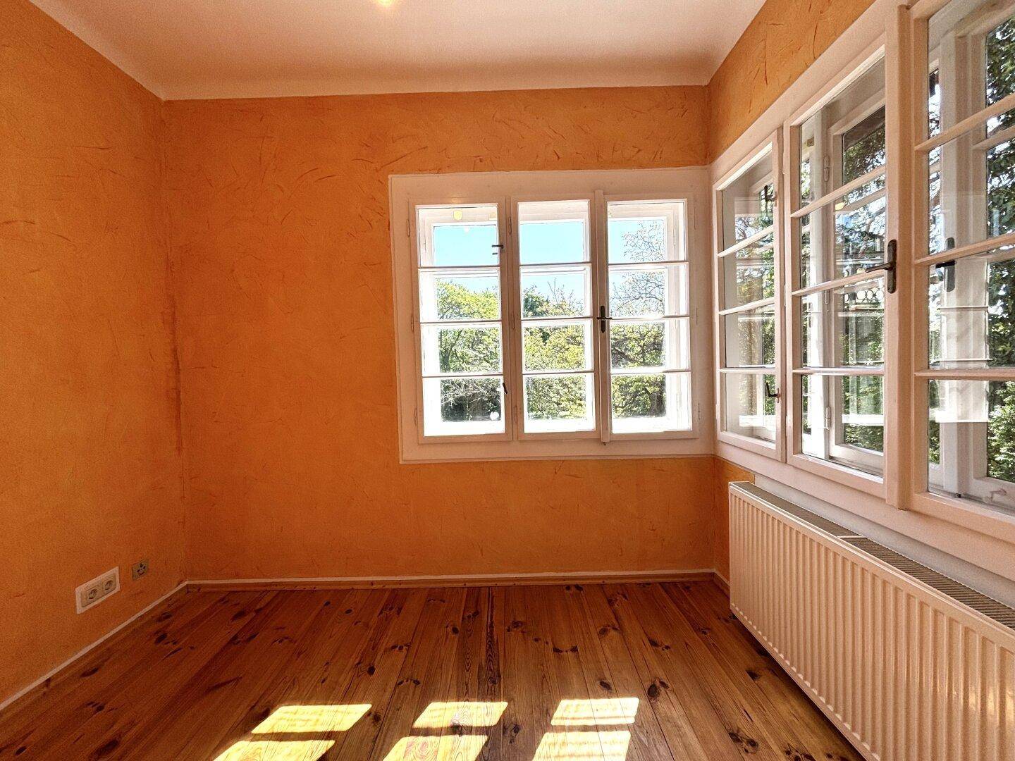 Lichtdurchflutetes Zimmer mit Holzdielenboden, gelben Wänden und großen Fenstern zum Garten.