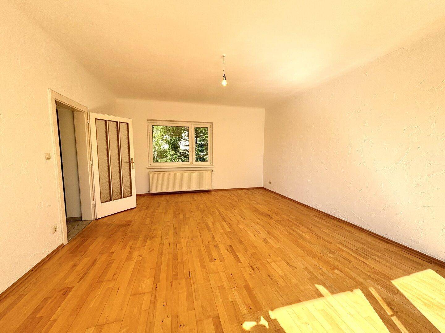 Geräumiges Wohnzimmer mit Parkettboden, weißen Wänden und einem Fenster mit Blick ins Grüne.