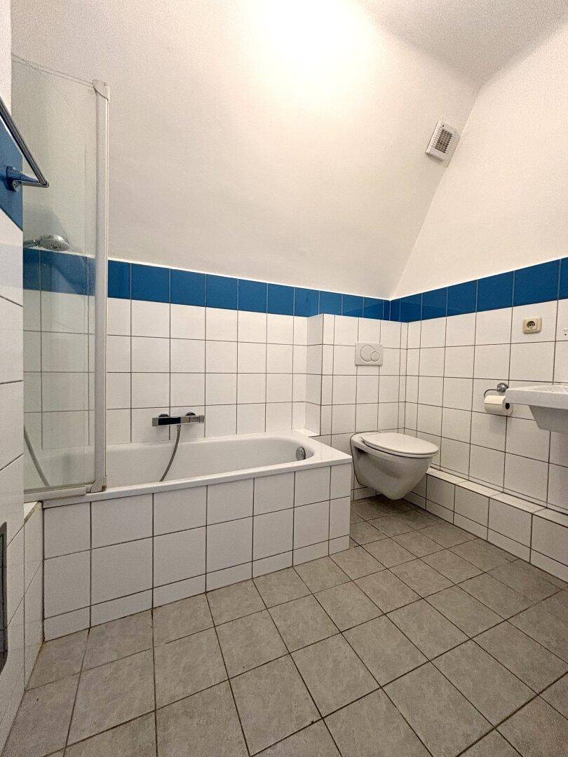 Badezimmer mit Badewanne, WC, Waschbecken und weißen Fliesen mit blauer Akzentlinie.