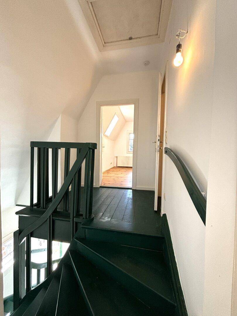 Treppenhaus mit dunkelgrün gestrichener Holztreppe und Zugang zu weiteren Räumen im Dachgeschoss.