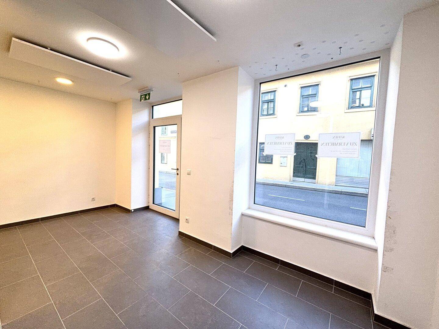 Leerer Ladenraum im Erdgeschoss mit großen Schaufenstern zur Straße und Fliesenboden.