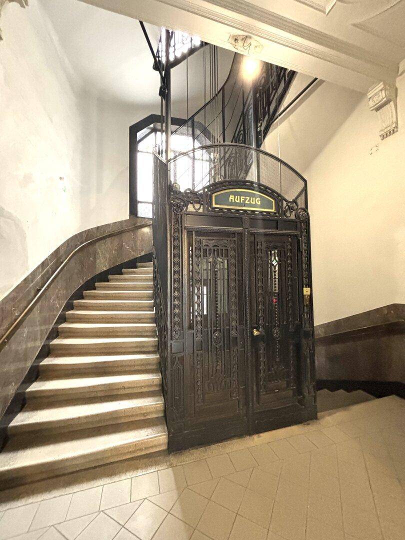 Historisches Treppenhaus mit Marmorstufen und einem kunstvoll verzierten Aufzug, der den Charme des Gebäudes unterstreicht.