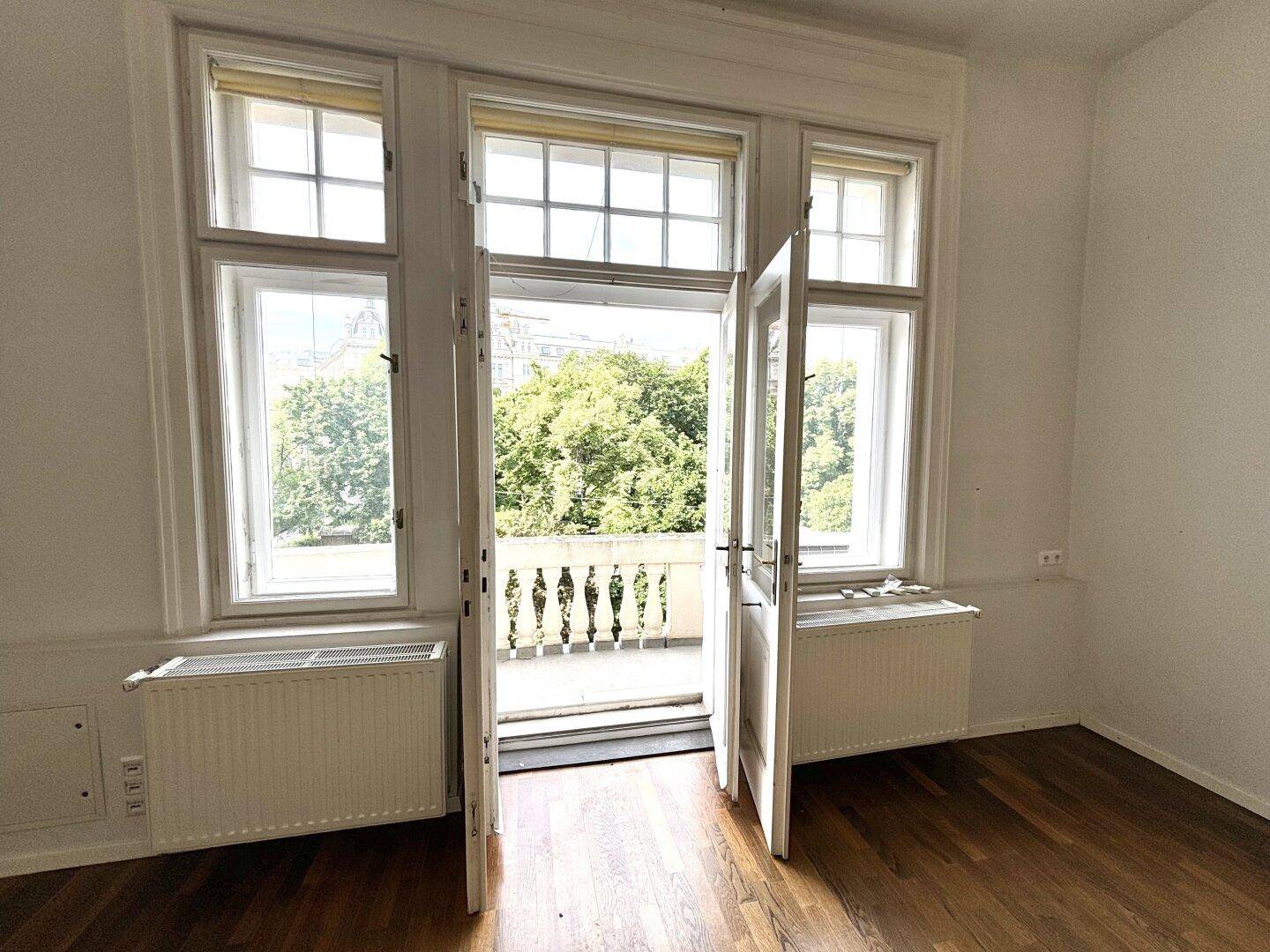 Innenansicht eines Raumes mit Holzboden und geöffneten Türen, die den Zugang zum Balkon ermöglichen.