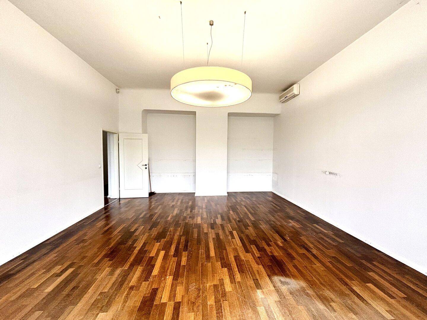 Geräumiger, leerer Raum mit Holzboden und einem Durchgang zu einem weiteren Bereich, ideal für Büros.