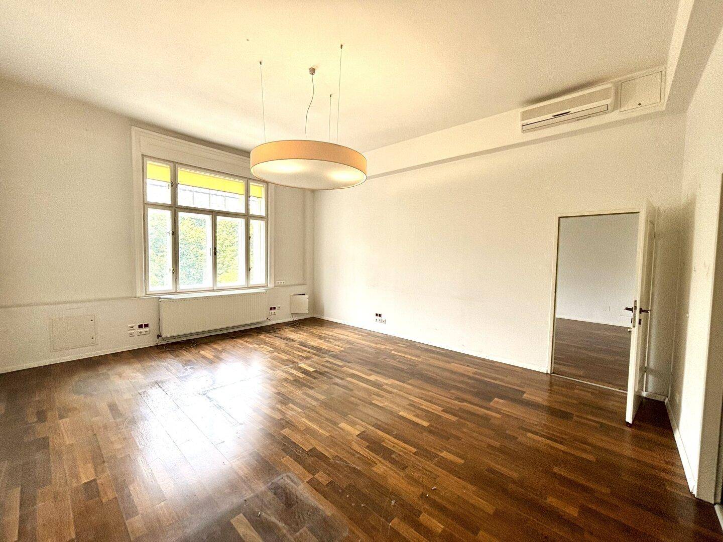 Geräumiger, leerer Raum mit Holzboden, großen Fenstern und Klimaanlage, ideal für Bürozwecke.