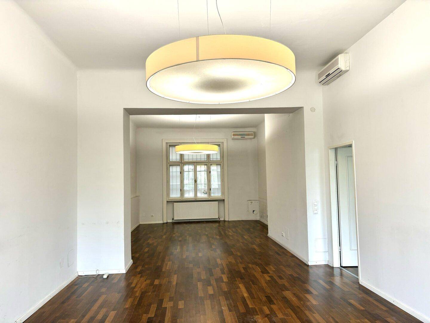 Geräumiger, heller Raum mit Holzboden und einem Durchgang zu einem weiteren Bereich, ideal für Büros.