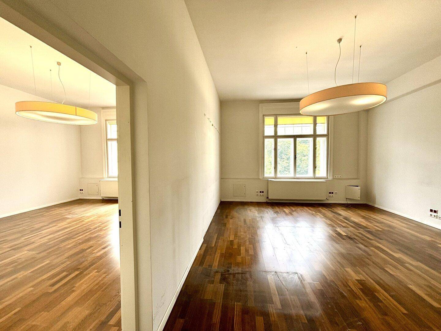 Zwei miteinander verbundene, helle Räume mit Holzboden und großen Fenstern, die viel Licht hereinlassen.