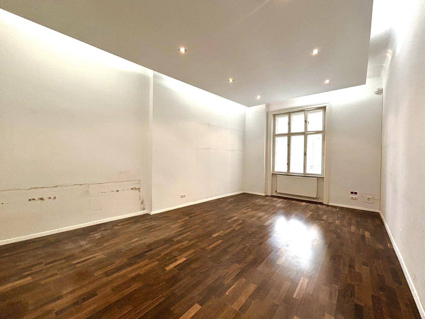Heller, leerer Raum mit Holzboden, Einbaudeckenleuchten und einem großen Fenster, das Tageslicht spendet.