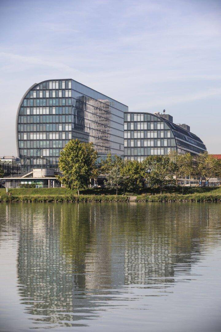 Modernes Gebäude mit geschwungener Glasfassade, das sich im ruhigen Wasser eines Flusses spiegelt, umgeben von Bäumen.