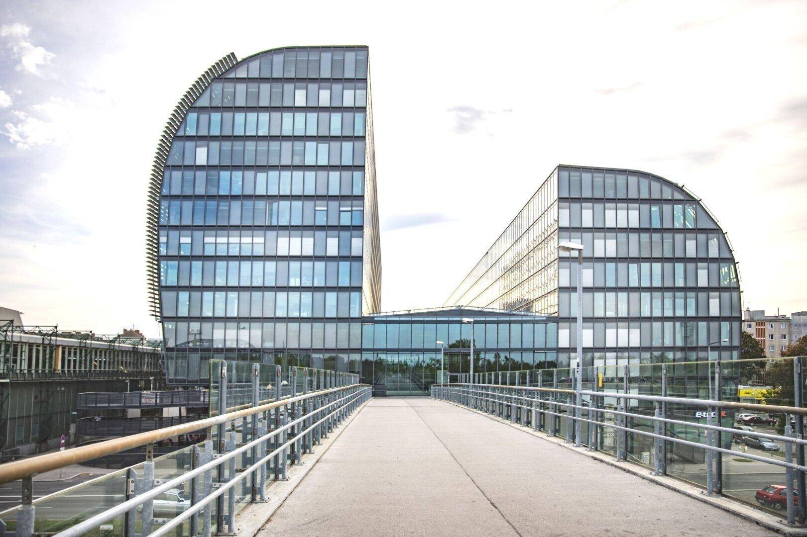 Ansicht des modernen Bürogebäudes mit geschwungener Glasfassade, aufgenommen von einer erhöhten Fußgängerbrücke.