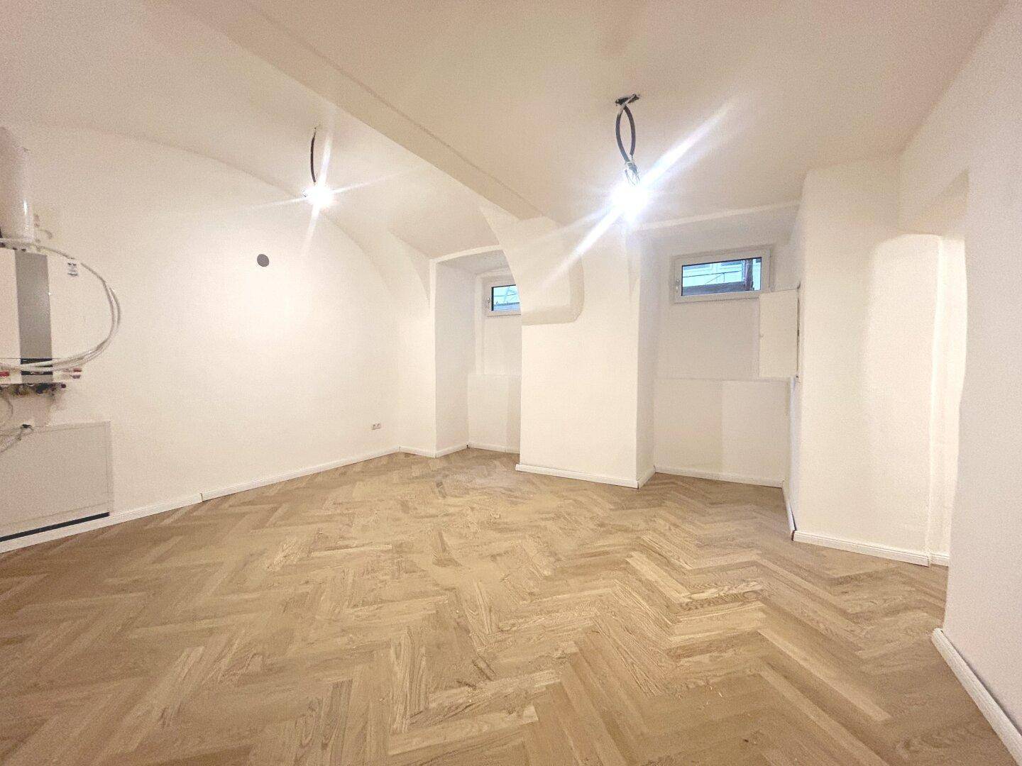 Leerer Raum mit Fischgrätparkett, weißen Wänden und sichtbaren Installationen an der Seite.