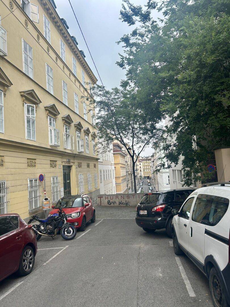Blick auf eine belebte Straße mit parkenden Autos, Bäumen und historischen Gebäuden im Hintergrund.