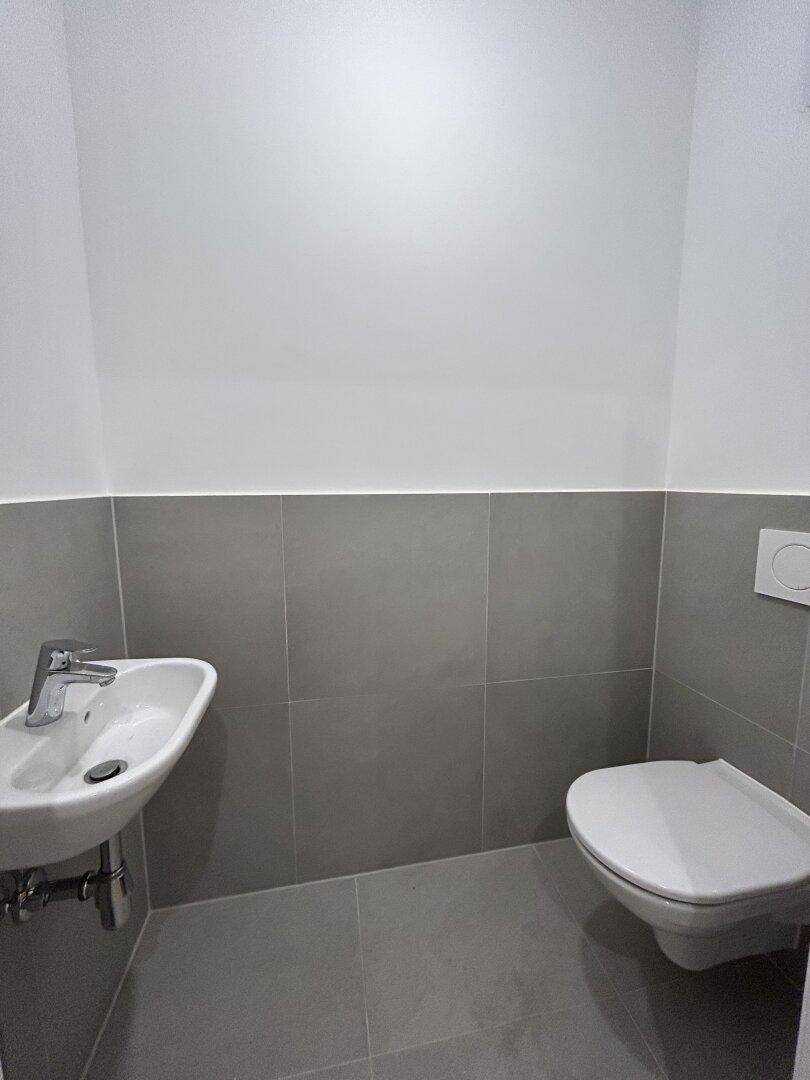 Moderne Gästetoilette mit grauen Wand- und Bodenfliesen, wandhängendem WC und kleinem Waschbecken.