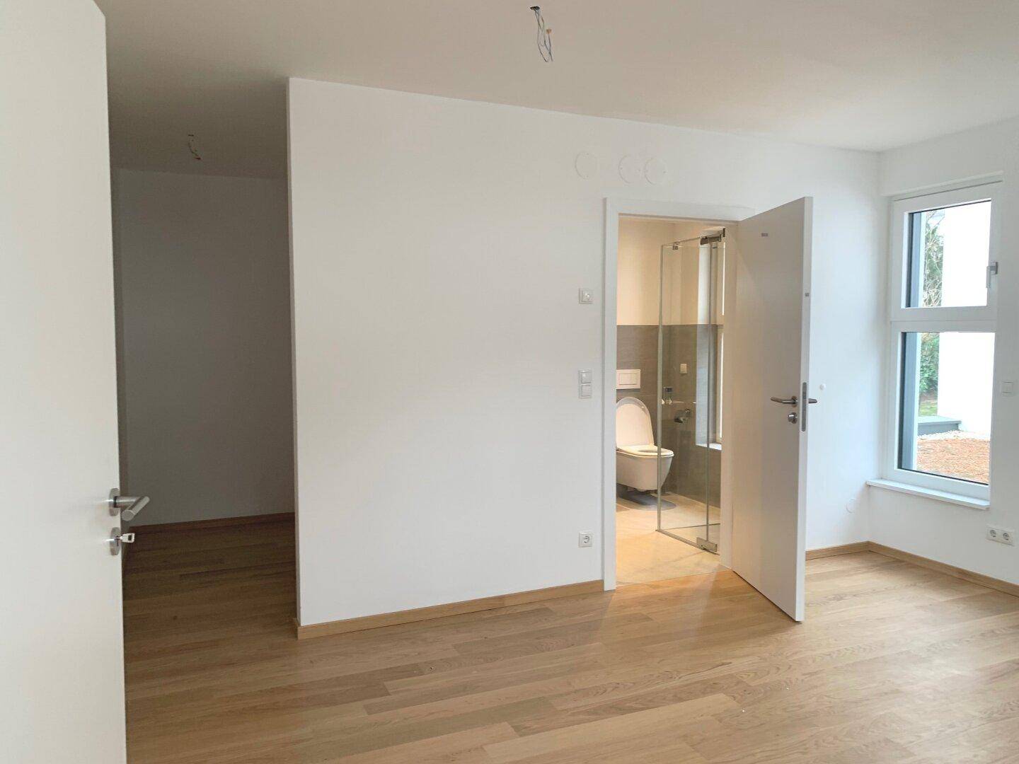 Leeres Schlafzimmer mit Holzboden und offener Tür zu einem modernen Badezimmer.