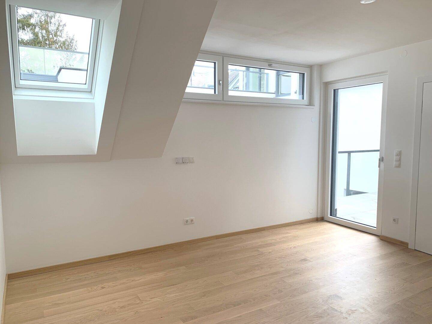 Leeres Schlafzimmer im Dachgeschoss mit Holzboden, Dachfenstern und Zugang zu einem Balkon.