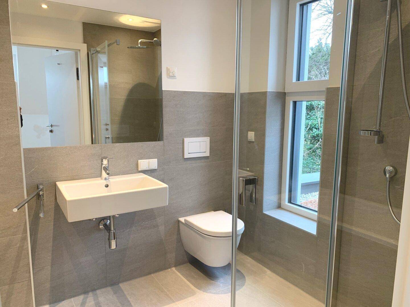 Modernes Badezimmer mit grauen Fliesen, wandhängendem WC und ebenerdiger Dusche.