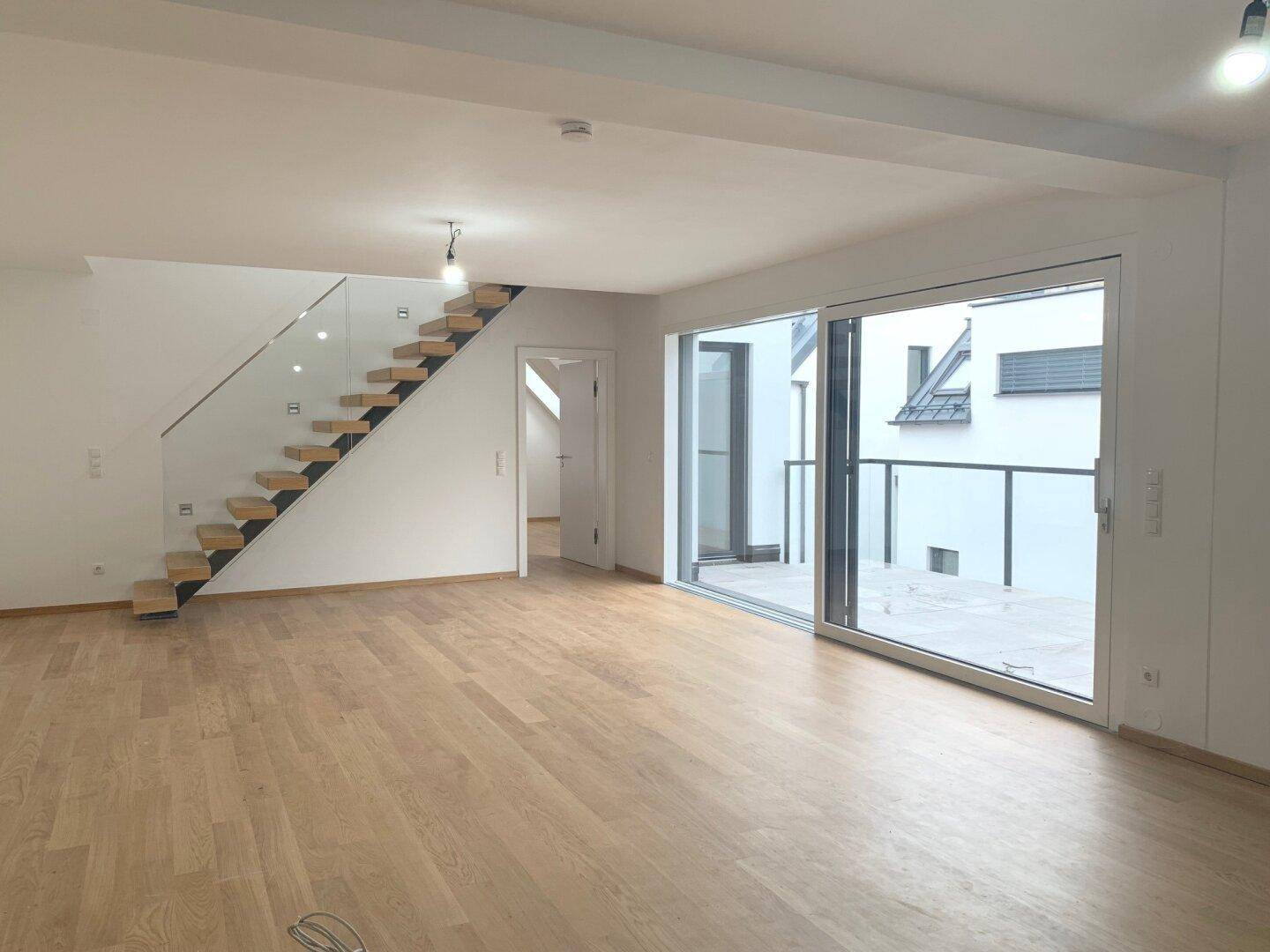 Geräumiger, leerer Wohnbereich mit Holzboden, offener Treppe und großen Schiebetüren zur Terrasse.