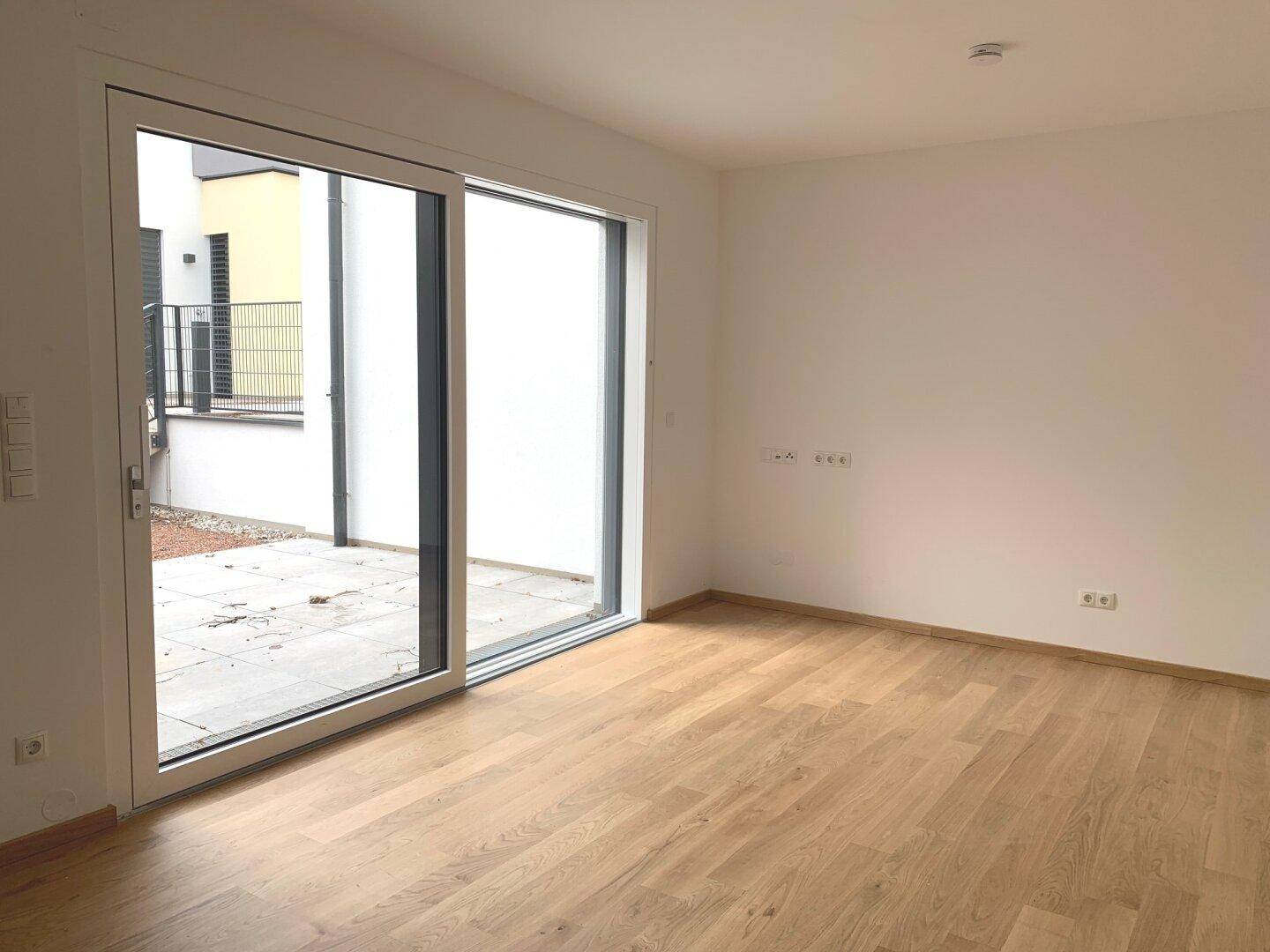 Leeres Zimmer mit Holzboden und Schiebetür zu einem kleinen Außenbereich.