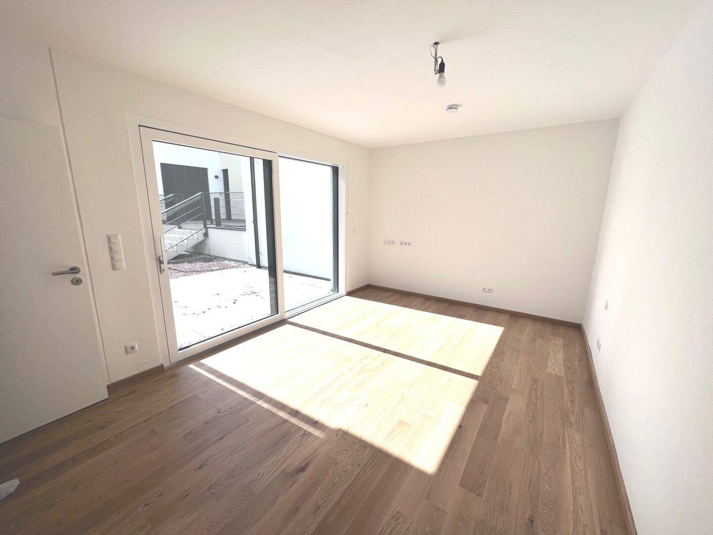 Leeres Zimmer mit Holzboden und Schiebetür zu einem kleinen Außenbereich.