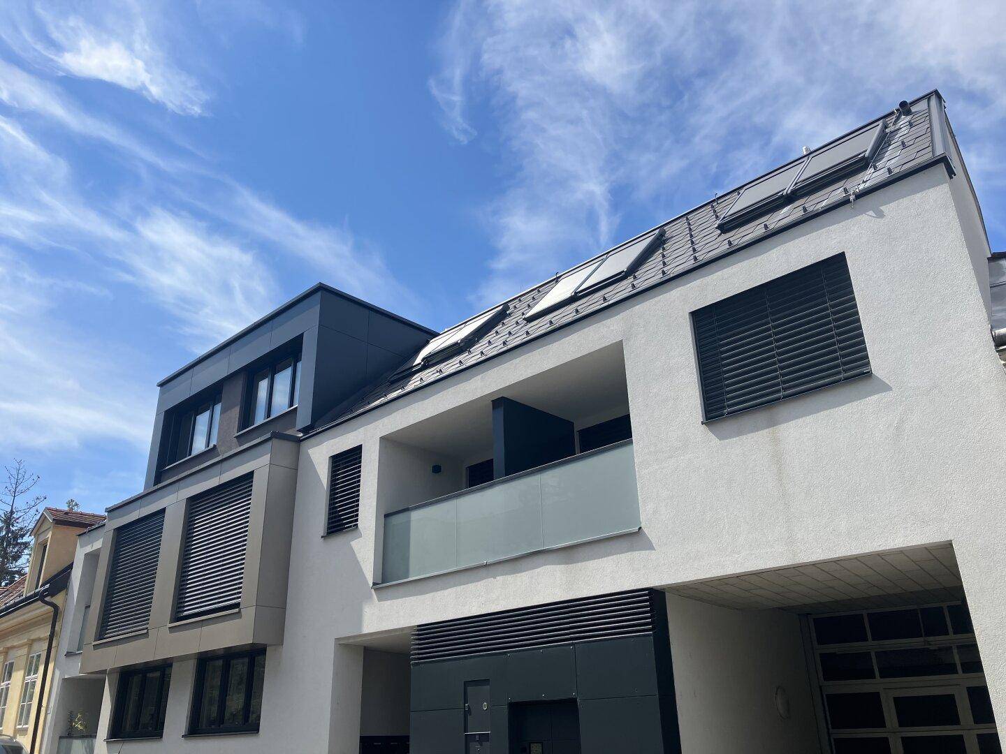 Moderne Gebäudefassade mit Balkonen, großen Fenstern und Dachfenstern unter blauem Himmel.