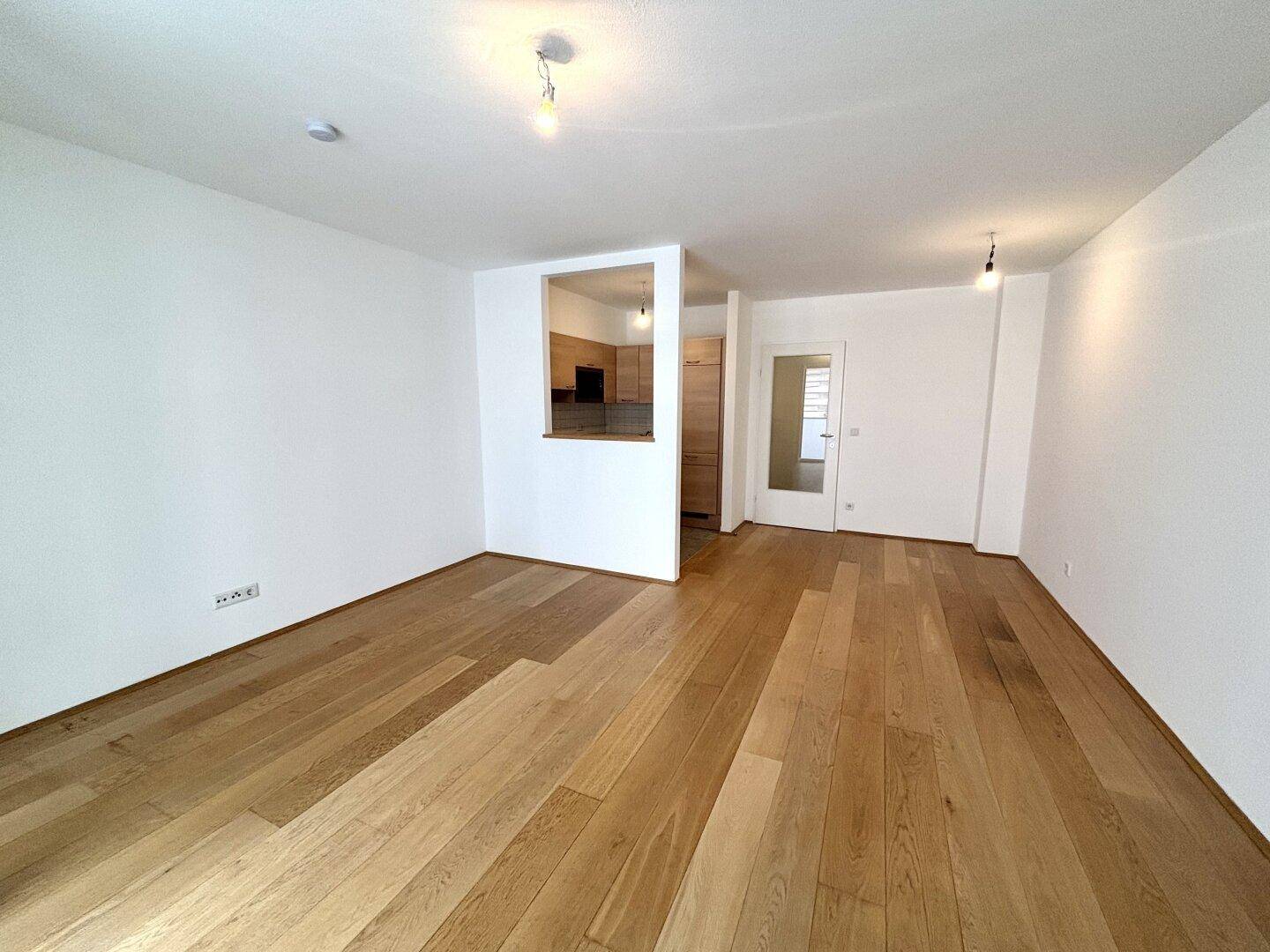 Heller, leerer Wohnbereich mit Holzboden und offener Verbindung zur Küche.