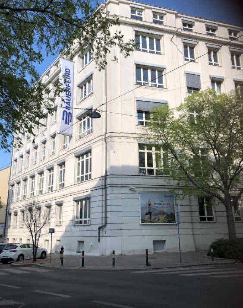 Ansicht der Gebäudeecke an einer Straße, umgeben von Bäumen und mit einem Werbebanner an der Fassade.