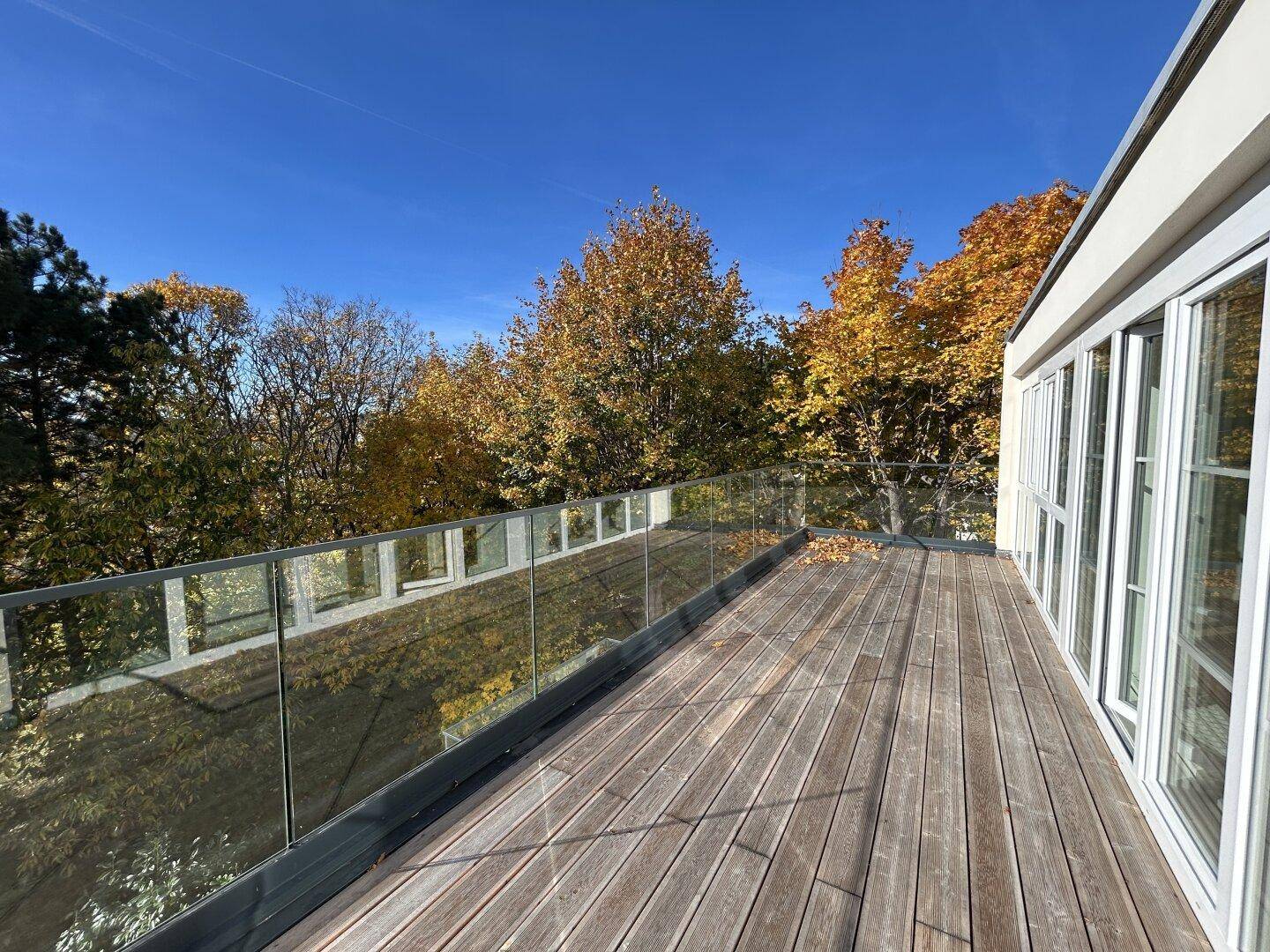 Weitläufige Holzterrasse mit Glasgeländer und Blick auf herbstliche Bäume unter blauem Himmel.