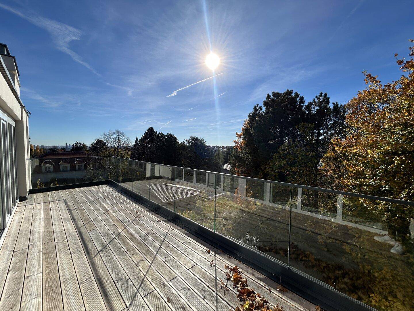 Sonnige Holzterrasse mit Glasgeländer und weitem Blick über die Landschaft und herbstliche Bäume.