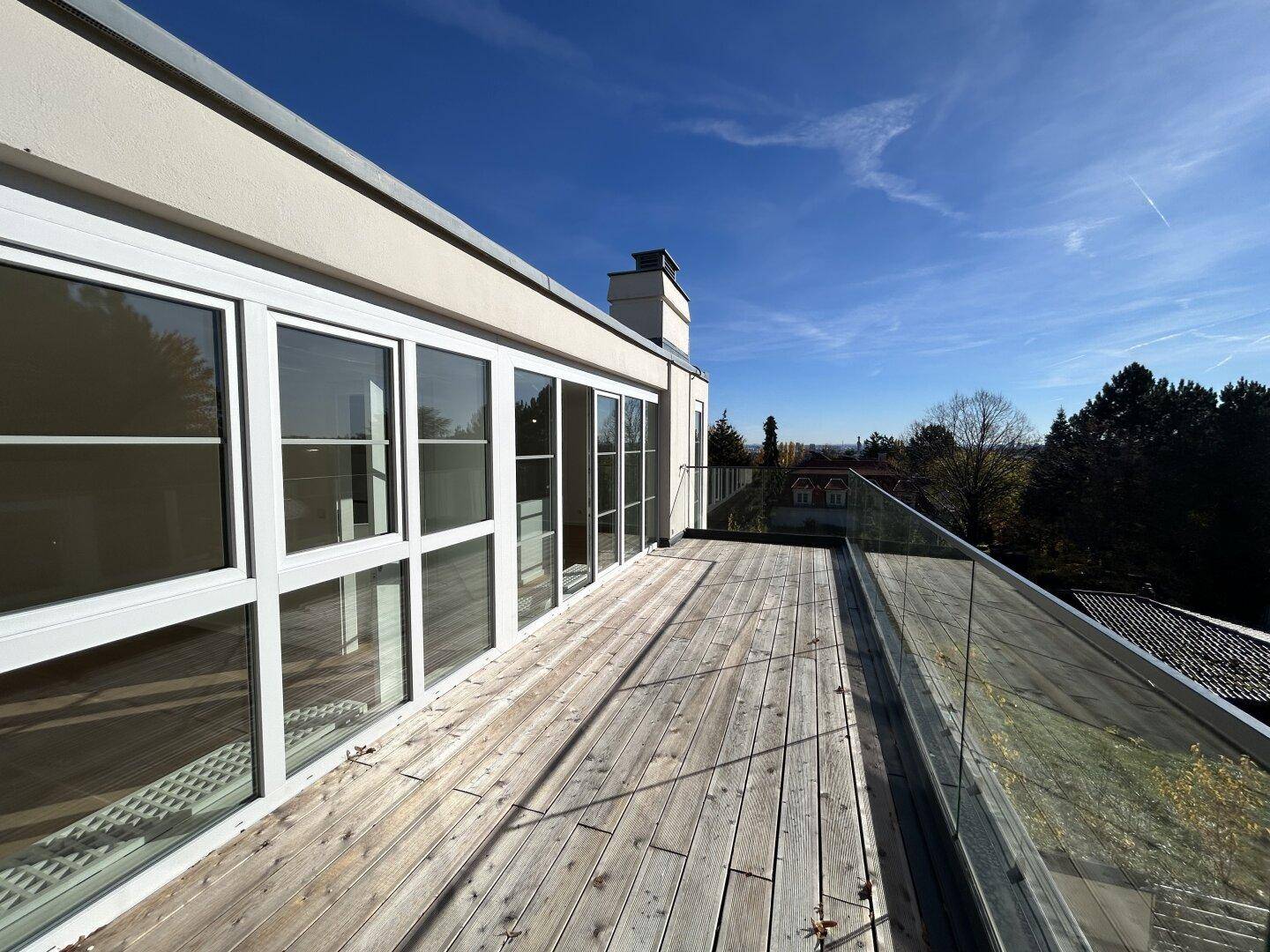 Geräumige Holzterrasse entlang der Gebäudefassade mit großen Fenstern und weitem Ausblick.