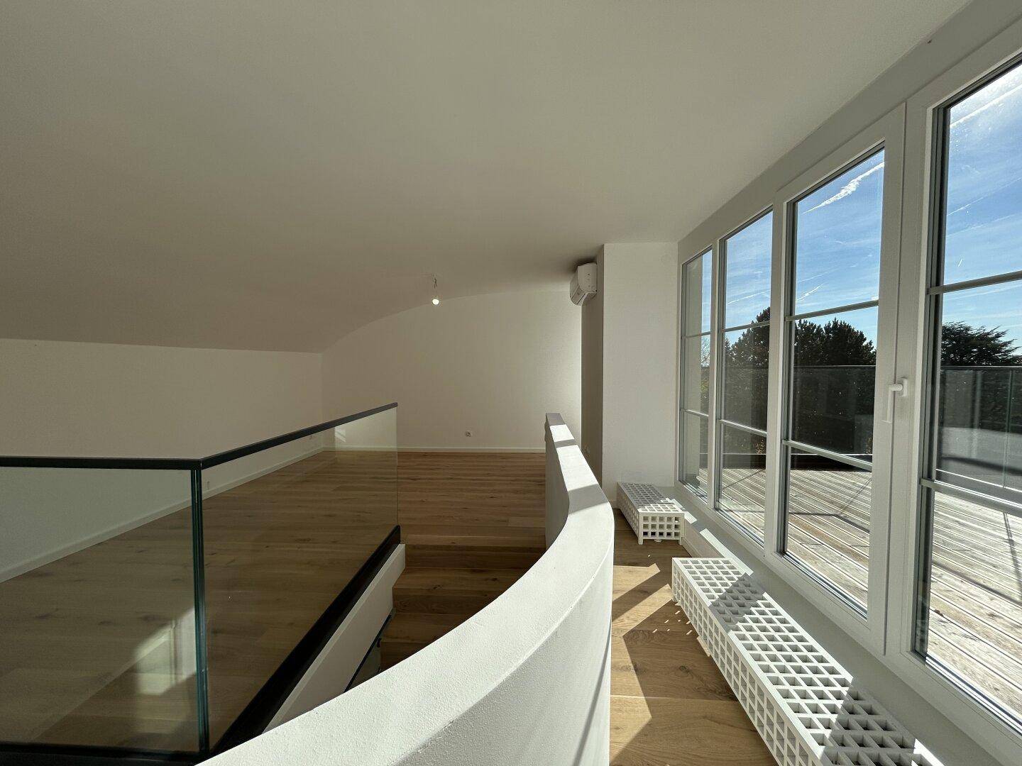 Heller offener Wohnbereich im Obergeschoss mit Holzboden, Glasgeländer und Zugang zur Terrasse.