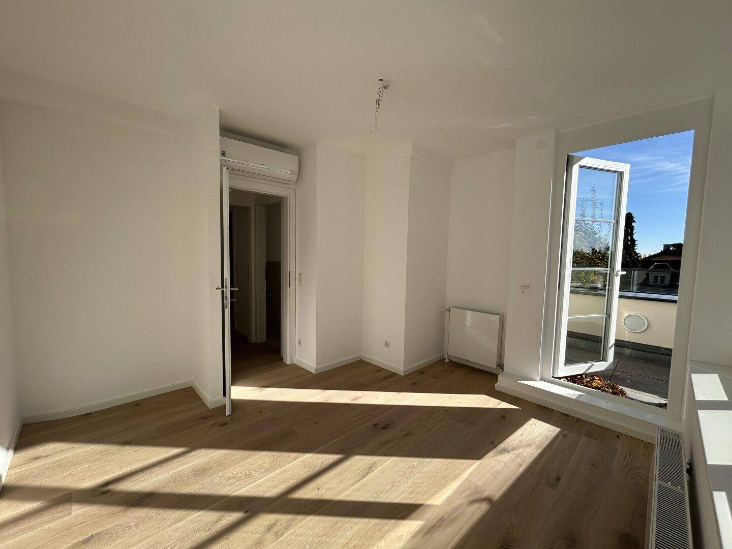 Heller Raum mit Holzboden und einer Tür, die zu einem Balkon oder einer Terrasse führt.