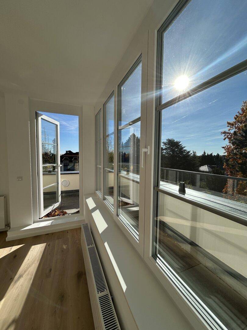 Lichtdurchfluteter Raum mit großen Fenstern, Holzboden und Zugang zu einem sonnigen Außenbereich.