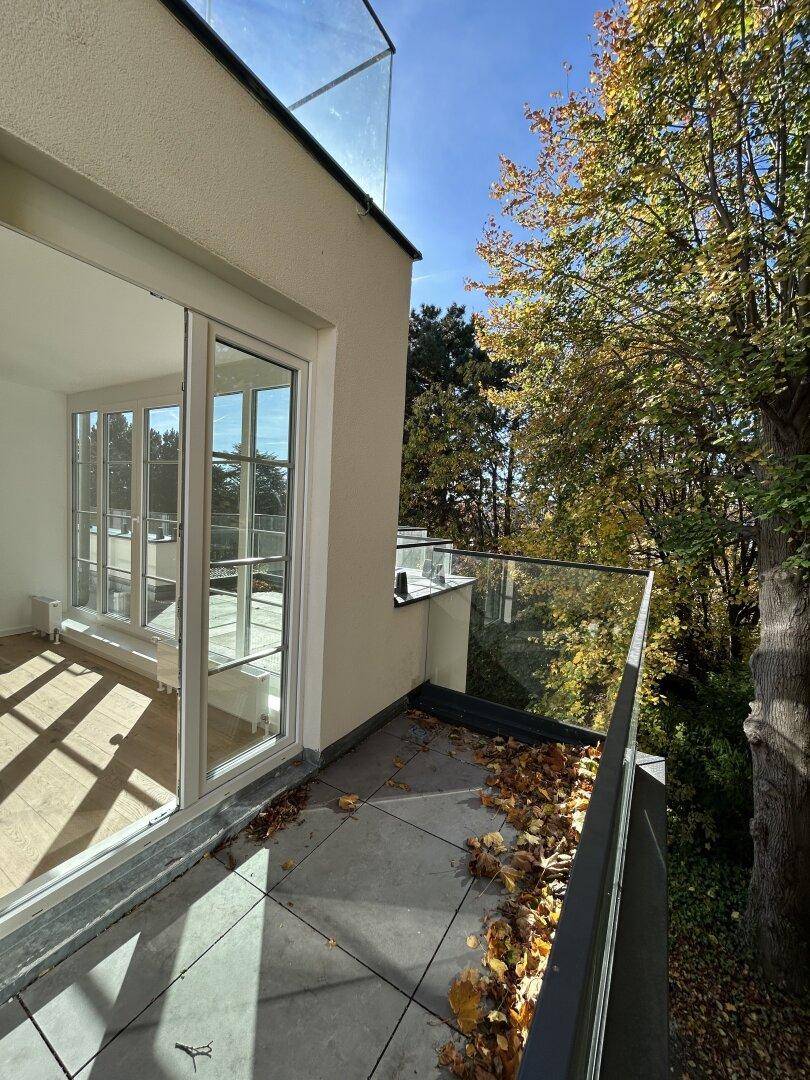Kleiner Balkon mit Glasgeländer und Zugang zum Innenraum, umgeben von herbstlichem Laub.