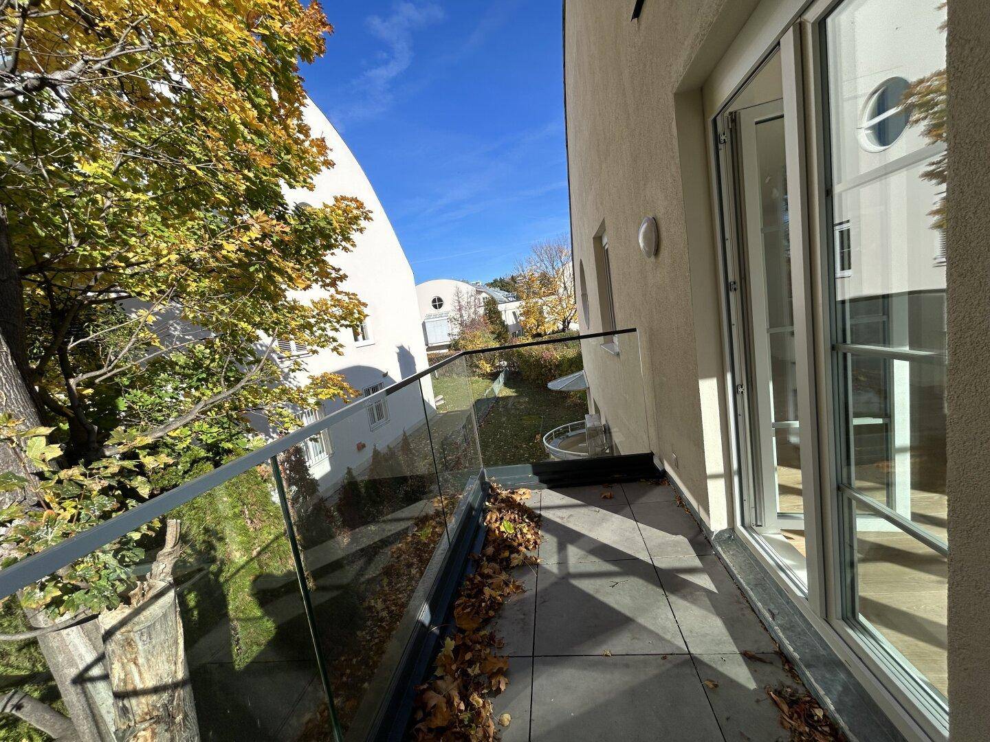 Kleiner Balkon mit Glasgeländer und Blick auf die grüne Umgebung und andere Gebäude.