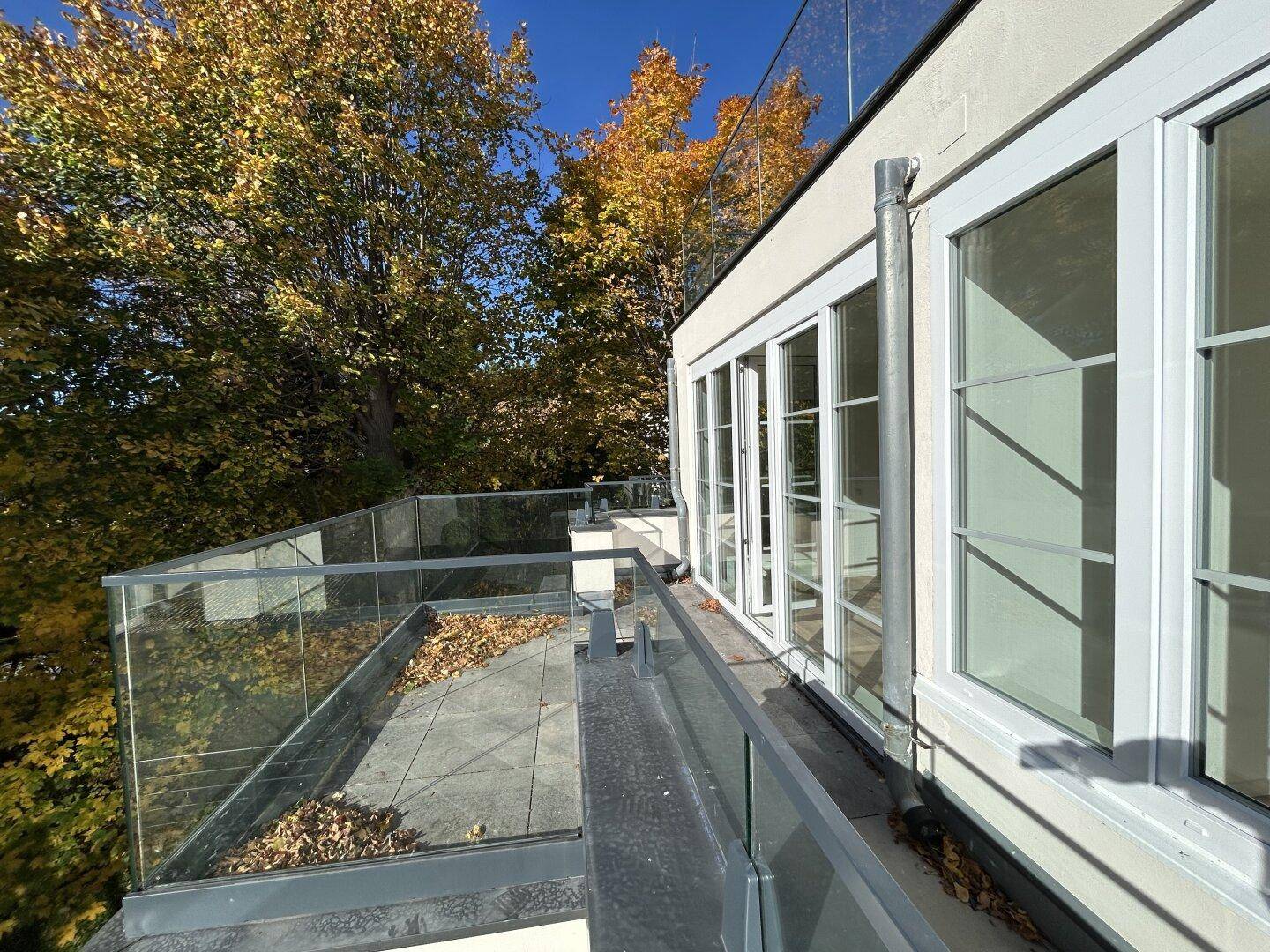 Geräumige Terrasse mit Glasgeländer und Blick auf herbstliche Bäume unter blauem Himmel.