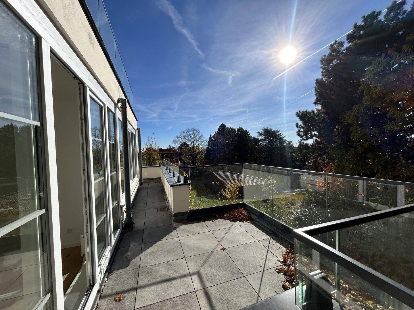 Sonnige Terrasse mit Fliesenboden und Glasgeländer, umgeben von grünen Pflanzen.