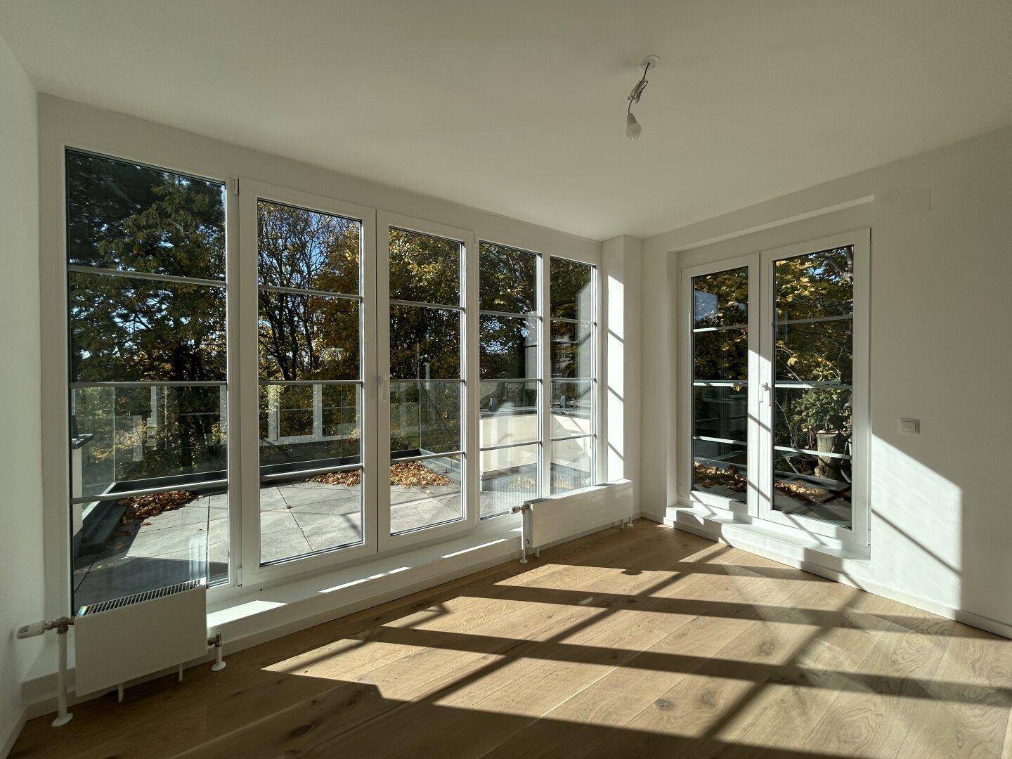 Heller Raum mit Holzboden und großen Fenstern, die Zugang zu einer Terrasse bieten.