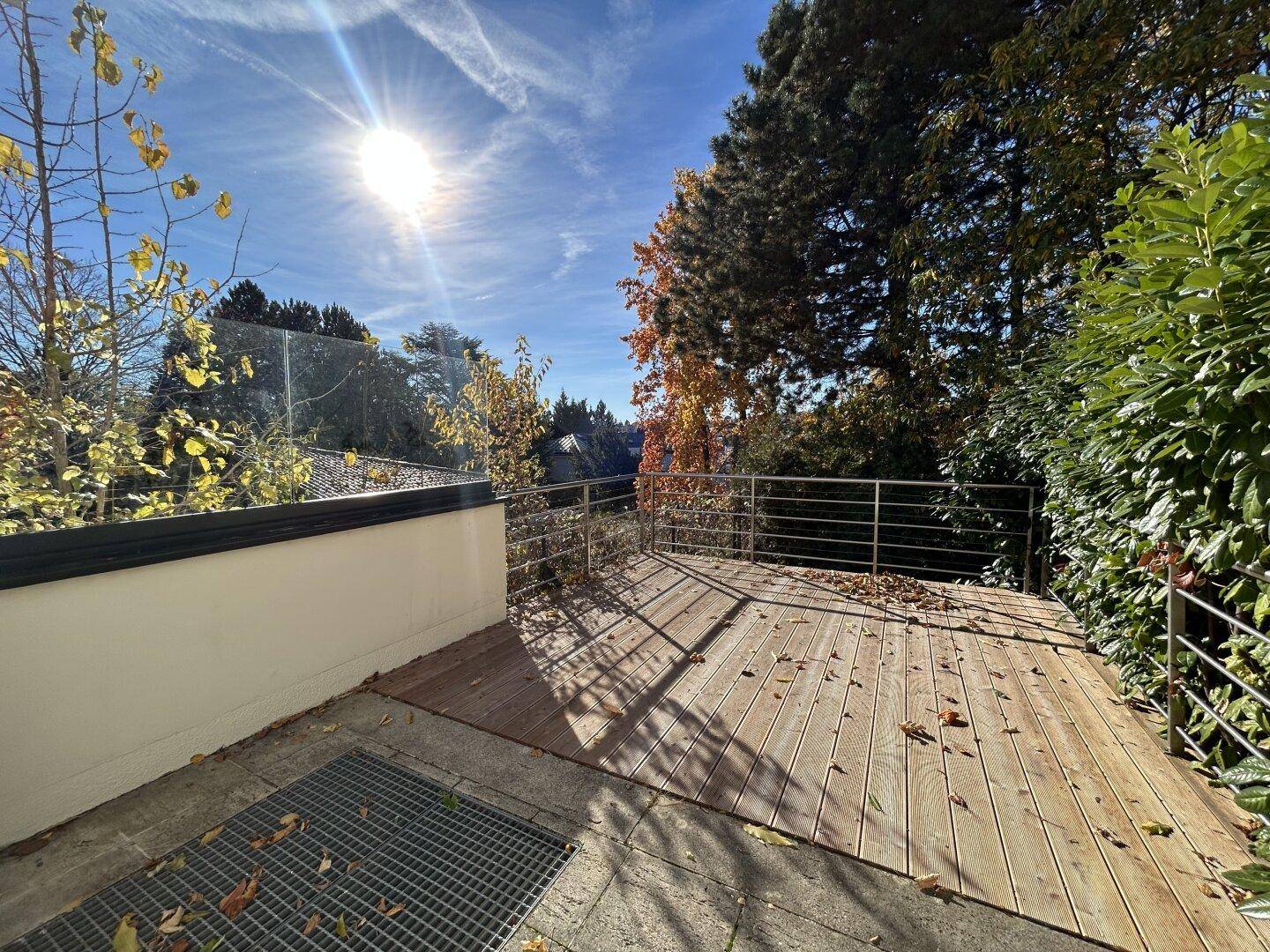 Sonnige Holzterrasse mit Glasgeländer und Blick ins Grüne, umgeben von Bäumen.