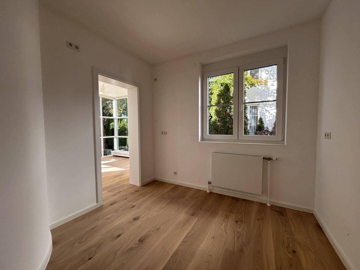 Leerer Raum mit Holzboden, Fenster und Zugang zu einem weiteren hellen Zimmer.